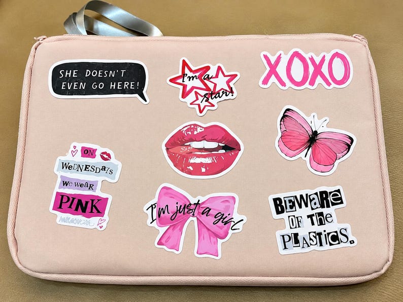 May include: A light pink rectangular laptop sleeve decorated with various stickers including a pink butterfly, red lips, a pink bow, and text phrases such as "She doesn't even go here," "XOXO," "On Wednesdays we wear pink," "I'm just a girl," and "Beware of the plastics."
