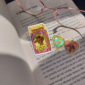 May include: Three stickers on a book page. One is a matchbox with a dachshund wearing heart-shaped sunglasses, the text "Long Burn". Two heart-shaped lockets with dachshunds. Rose gold glasses rest on the page.