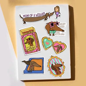 May include: A collection of colorful stickers featuring dachshund dogs in various poses. One sticker reads "MOM OF A WIENER", another says "SHORT LEGS BIG ATTITUDE!" and another says "LONG BUM". The stickers are on a white background.