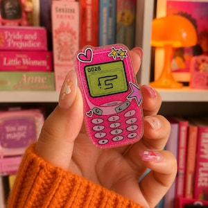May include: A glittery pink acrylic pin shaped like a retro mobile phone. The screen displays the text 0028 and a simple line drawing. The pin features a numeric keypad, a pink heart icon, two yellow stars, and a purple star accent on the side.