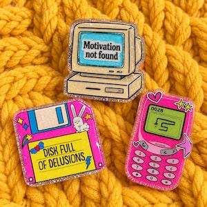 Puede incluir: Tres pines de acrílico con tecnología retro: un monitor de computadora beige que muestra "Motivation not found", un disquete rosa con la etiqueta "DISK FULL OF DELUSIONS" y un gráfico de conejo, y un teléfono móvil rosa con una pantalla de juego.