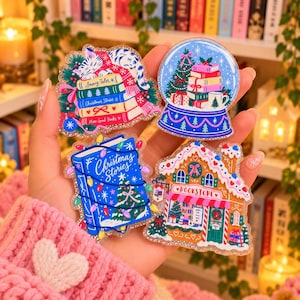 May include: Four glittery acrylic stickers featuring holiday themes: a cat sleeping on books labelled Snowy Tales and Christmas Stories, a snow globe with a tree and books, a blue book titled Christmas Stories, and a gingerbread bookstore.