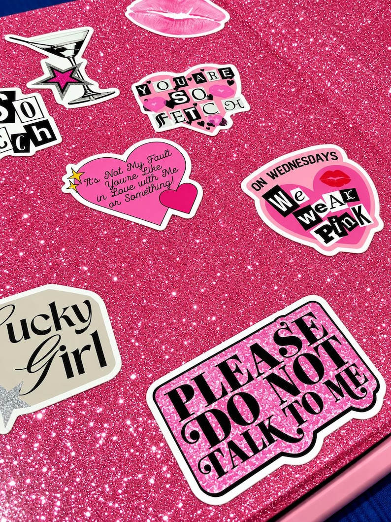 May include: A bright pink glittery surface covered in various stickers including a martini glass, a pair of lips, and heart-shaped designs with text such as "You are so fetch," "It's not my fault you're like in love with me," and "Please do not talk to me."