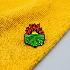 May include: A green enamel pin shaped like a dumpster on fire with red and orange flames. The text DUMPSTER FIRE RESPONSE TEAM and TEAM LEADER is printed on the front in black, accompanied by three yellow stars on the side of the dumpster.