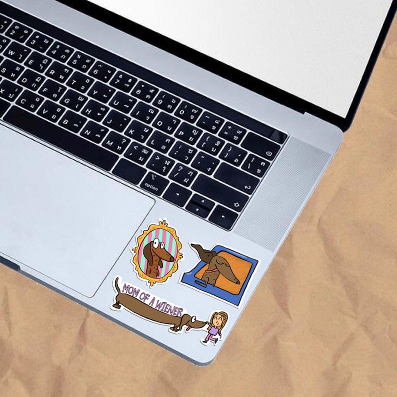 May include: A laptop with three dachshund-themed stickers. One sticker features a dachshund in a gold frame, another shows a dachshund in a blue box, and the third says "MOM OF A WIENER" with a cartoon dachshund and girl.