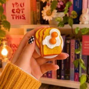 May include: A circular phone grip featuring a miniature three-dimensional sculpture of a slice of golden-brown toasted bread topped with a fried egg that has two bright orange yolks. The toast has a textured crust and a smooth, glossy finish.