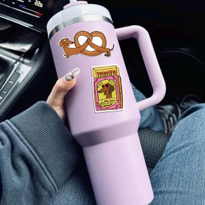 May include: A large, light purple insulated tumbler with a white lid and handle. The tumbler is decorated with two stickers: one of a dachshund pretzel and another of a dachshund wearing sunglasses with the text "Long Burn".