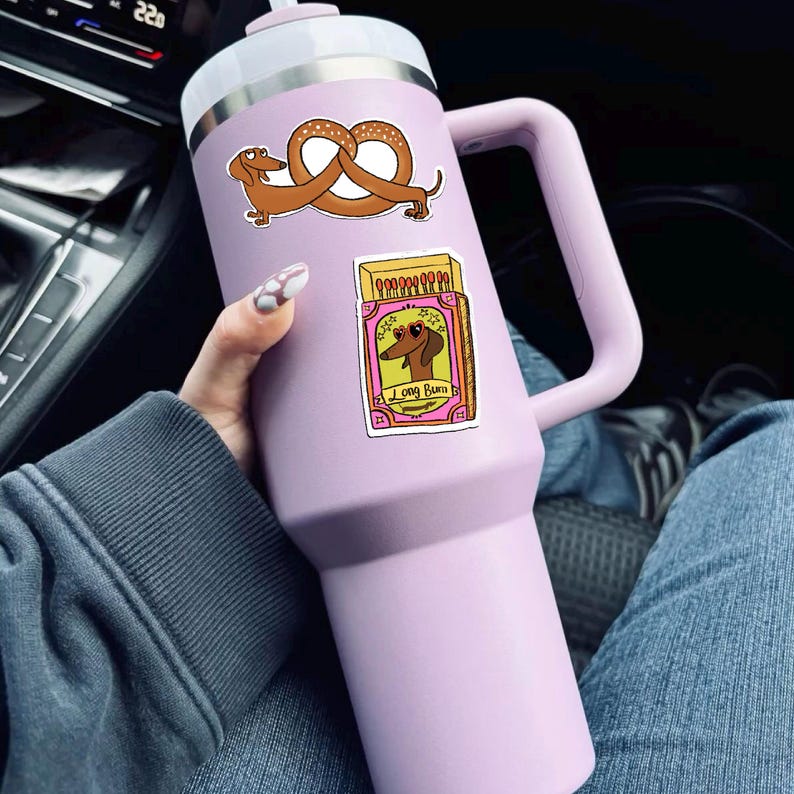 May include: A large, light purple insulated tumbler with a white lid and handle. The tumbler is decorated with two stickers: one of a dachshund pretzel and another of a dachshund wearing sunglasses with the text "Long Burn".