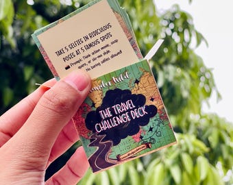 May include: A small rectangular card deck titled The Travel Challenge Deck with a map background. One card shows the text: Take 5 selfies in ridiculous poses at 5 famous spots. Prompt: Think action movie, soap opera, or slo-mo style. No boring selfies allowed!