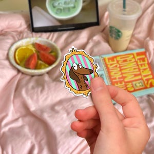 May include: A colorful sticker featuring a cartoon dachshund dog inside a decorative oval frame with pink and green stripes. The frame has a gold border. The sticker is held in front of a book titled "The House on Mango Street".