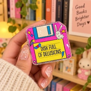 May include: A square glittery pin shaped like a floppy disk with a bright pink and yellow background. It features a white rabbit illustration, a blue heart with wings, and the black text DISK FULL OF DELUSIONS with a blue lightning bolt icon.