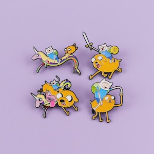 May include: Four enamel pins featuring characters from the animated series Adventure Time. Each pin depicts Finn and Jake in various poses, riding a unicorn, a rainbow, and Jake himself. The pins are set against a purple background.