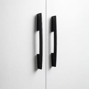 May include: Two black wooden cabinet pulls are mounted on a white cabinet door. The pulls have a rectangular shape with a curved design and a cutout in the center. The hardware is made of wood and has a dark finish.