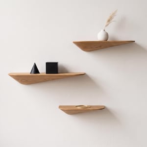 Könnte beinhalten: Drei wandmontierte Holzregale mit einem sich verjüngenden, geschwungenen Unterseitendesign. Das oberste Regal trägt eine weiße Vase mit einer getrockneten Pflanze. Das mittlere Regal trägt einen schwarzen Kegel und einen schwarzen Würfel.