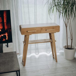 Puede incluir: Una consola de madera clara con dos cajones y un estante inferior. La mesa tiene un diseño moderno de mediados de siglo con patas anguladas y tiradores de madera. Una planta en maceta está a la derecha de la mesa.