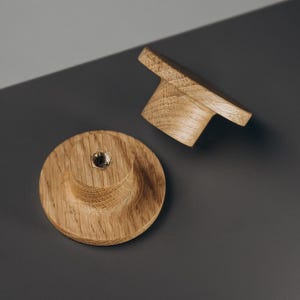 Peut inclure: Deux boutons de meuble en bois avec une finition naturelle. L'un est rond avec un centre sur&eacute;lev&eacute; et un insert m&eacute;tallique. L'autre est rectangulaire avec une poign&eacute;e incurv&eacute;e. Les boutons sont en bois clair.