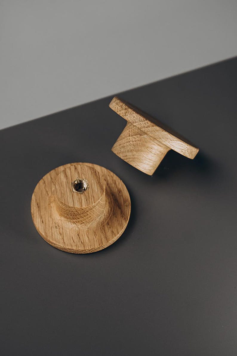 Peut inclure: Deux boutons de meuble en bois avec une finition naturelle. L'un est rond avec un centre sur&eacute;lev&eacute; et un insert m&eacute;tallique. L'autre est rectangulaire avec une poign&eacute;e incurv&eacute;e. Les boutons sont en bois clair.