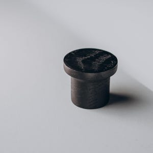 Peut inclure: Un bouton de meuble cylindrique en bois fonc&eacute; avec un dessus plat et circulaire. Le bouton est d'un noir charbon profond, et le grain du bois est visible sur la surface sup&eacute;rieure. Design simple et moderne, pour meubles ou armoires.