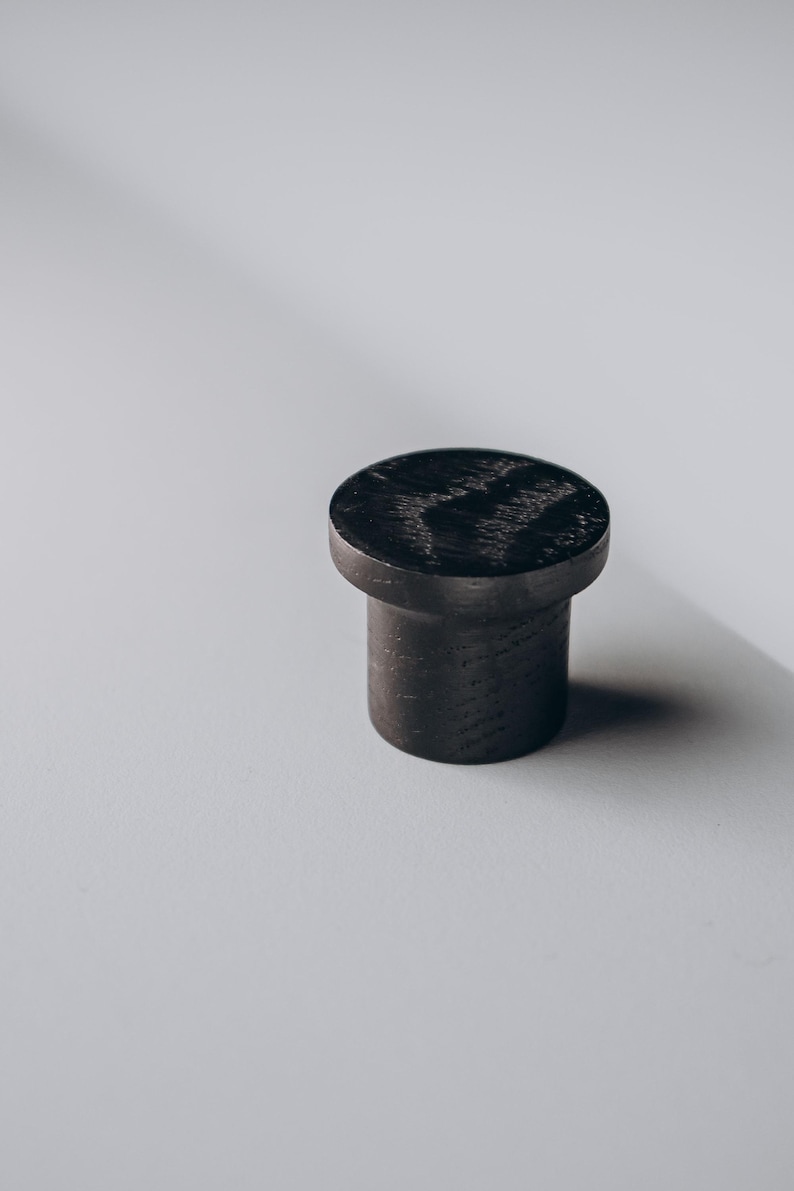 Peut inclure: Un bouton de meuble cylindrique en bois fonc&eacute; avec un dessus plat et circulaire. Le bouton est d'un noir charbon profond, et le grain du bois est visible sur la surface sup&eacute;rieure. Design simple et moderne, pour meubles ou armoires.