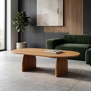 May include: An oval coffee table made of light-toned wood with a smooth surface and two curved, solid panel legs. A single rectangular book with a white cover rests flat on the center of the tabletop, which features a visible natural wood grain.
