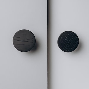 Peut inclure: Deux boutons de meuble ronds gris fonc&eacute; avec une texture de grain de bois sont mont&eacute;s sur une surface blanche. Les boutons sont espac&eacute;s uniform&eacute;ment et semblent &ecirc;tre en bois. Le fond est blanc uni.
