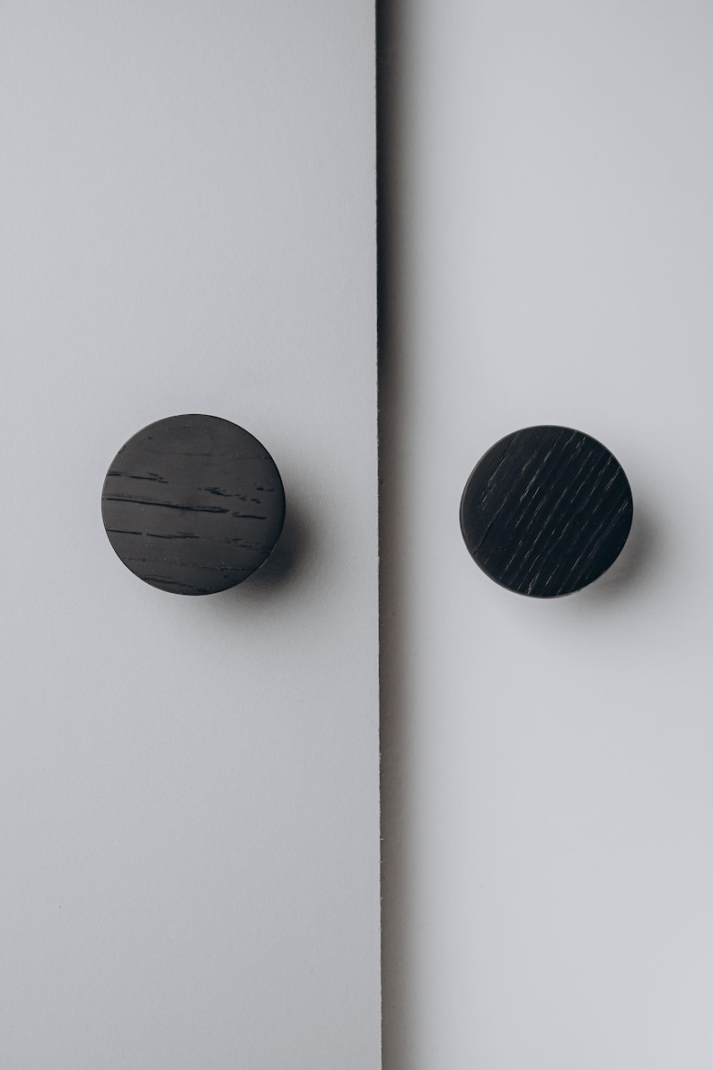 Peut inclure: Deux boutons de meuble ronds gris fonc&eacute; avec une texture de grain de bois sont mont&eacute;s sur une surface blanche. Les boutons sont espac&eacute;s uniform&eacute;ment et semblent &ecirc;tre en bois. Le fond est blanc uni.