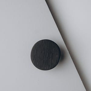 Peut inclure: Un bouton de meuble rond en bois gris fonc&eacute; avec une surface textur&eacute;e. Le bouton est pos&eacute; sur un fond blanc et gris, cr&eacute;ant une esth&eacute;tique minimaliste. Id&eacute;al pour les r&eacute;novations de meubles.