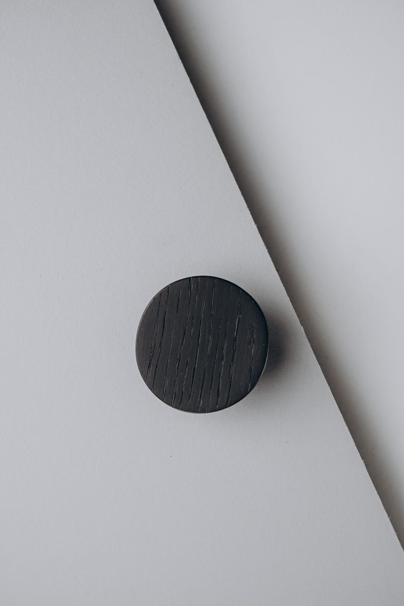 Peut inclure: Un bouton de meuble rond en bois gris fonc&eacute; avec une surface textur&eacute;e. Le bouton est pos&eacute; sur un fond blanc et gris, cr&eacute;ant une esth&eacute;tique minimaliste. Id&eacute;al pour les r&eacute;novations de meubles.