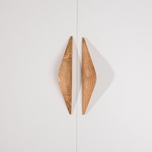 May include: Two vertical, diamond-shaped cabinet handles made of light brown wood with a visible natural grain pattern. The handles are mounted symmetrically on a smooth, solid white surface, creating a sharp, pointed silhouette with a curved profile.