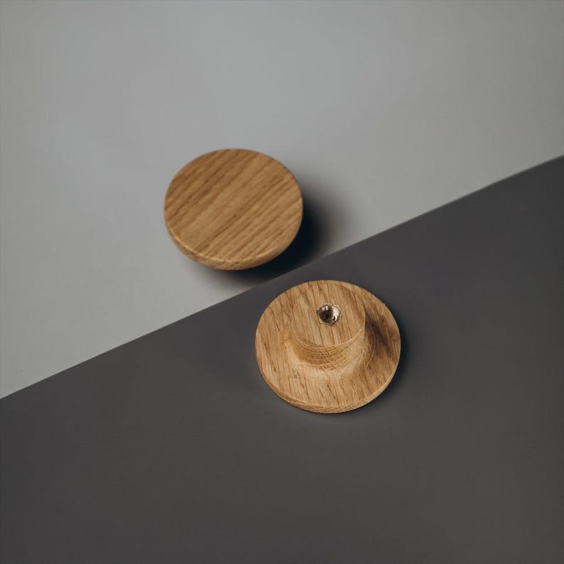 Peut inclure: Deux boutons de meuble en bois avec une finition naturelle vein&eacute;e. L'un est un simple disque, l'autre a une base arrondie et un insert m&eacute;tallique filet&eacute;. Les boutons sont pos&eacute;s sur un fond gris et gris clair.