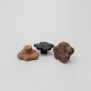 May include: Three flower-shaped wooden cabinet knobs. One is light brown, one is black, and the third is dark brown. The knobs are set against a white backdrop.