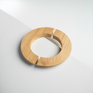 May include: A light brown wooden ring, with two gaps in the circle. The wood grain is visible. The ring is set against a white and light grey background.