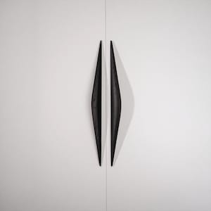 May include: Two long, vertical, black wooden cabinet handles with a tapered, spindle-like shape and visible wood grain texture. The handles are mounted side-by-side on a smooth, solid white surface, creating a symmetrical, elongated diamond profile.