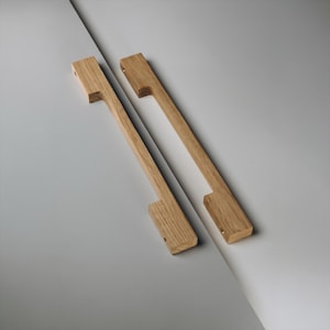 May include: Two wooden cabinet pulls with a rectangular design. The pulls are made of light-coloured wood and have a simple, modern aesthetic. Each pull has a rectangular handle with a cutout for gripping and is approximately 30 cm long.