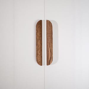 Puede incluir: Dos tiradores de armario de madera con la parte superior curvada y la inferior plana están montados en una puerta de armario blanca. Los tiradores son de madera marrón claro con un veteado visible.