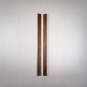 May include: Two vertical, dark brown wooden cabinet pulls are centered on a white cabinet door. The pulls are rectangular and have a wood grain texture. The cabinet door is plain and white.