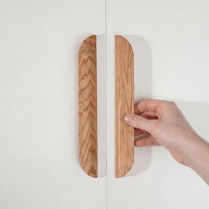 Könnte beinhalten: Zwei Holzschrankgriffe mit natürlicher Holzmaserung. Die Griffe sind hellbraun und haben oben und unten eine gebogene Halbmondform. Ein Griff wird von einer Hand gehalten.