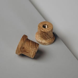 Peut inclure: Deux boutons de meuble en bois avec une finition naturelle vein&eacute;e. L'un des boutons est couch&eacute; sur le c&ocirc;t&eacute;, tandis que l'autre est debout. Les deux boutons ont un insert filet&eacute; pour une installation facile. Les boutons sont pos&eacute;s sur un fond gris.
