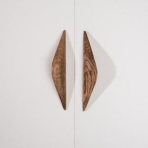 May include: Two wooden cabinet pulls with a diamond shape. The pulls are made of a light brown wood with a visible grain pattern. They are mounted on a white cabinet door. The pulls are approximately 6 inches long.