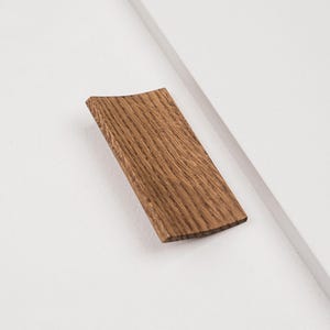 Pode incluir: Um puxador de armário retangular feito de madeira castanha com um padrão de grão natural visível. O puxador apresenta um perfil de superfície ligeiramente côncavo e curvo, montado numa superfície de painel branco com uma borda diagonal.