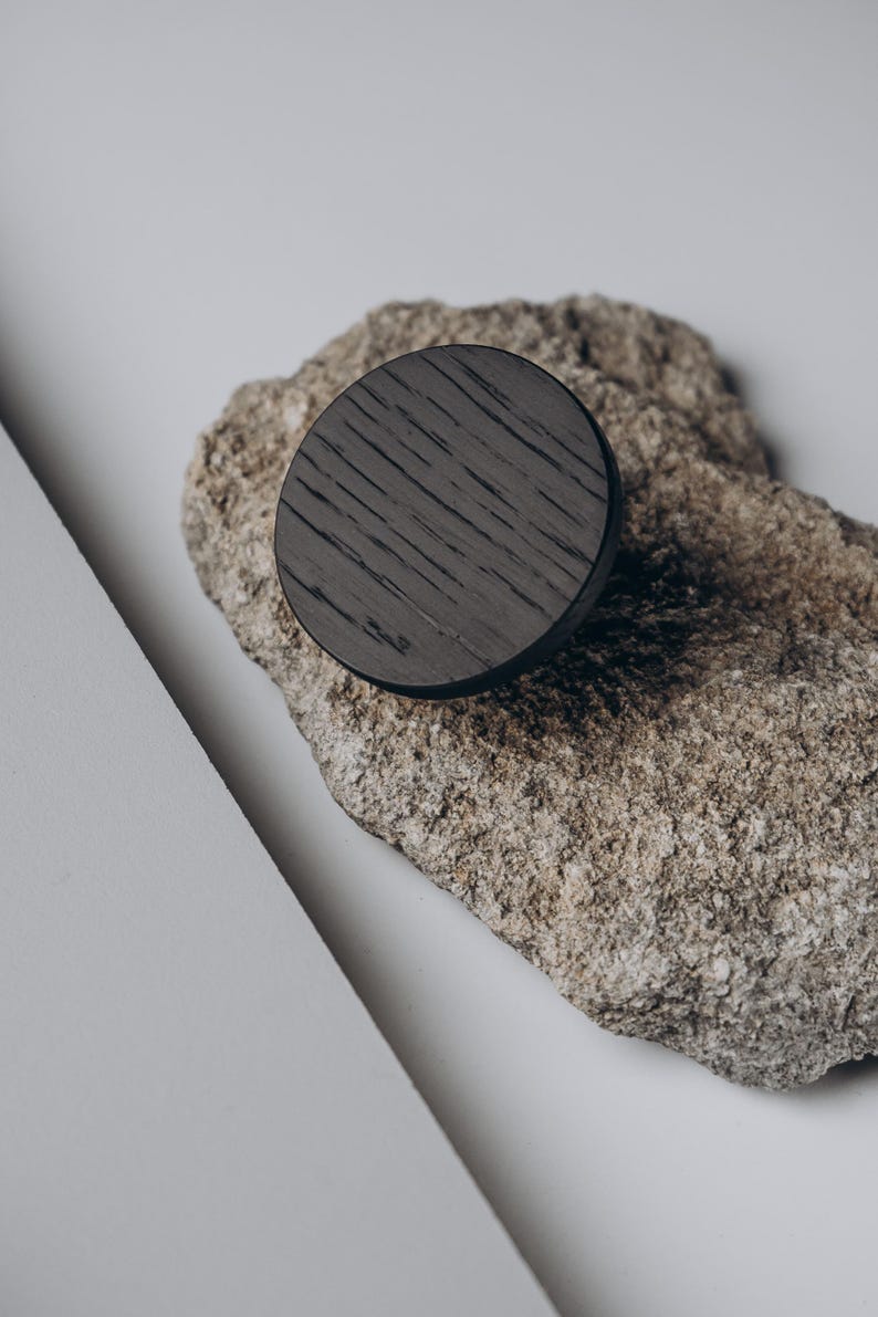 Peut inclure: Un objet rond en bois fonc&eacute; avec un grain visible est pos&eacute; sur une pierre rugueuse brun clair. La pi&egrave;ce en bois est de forme circulaire, peut-&ecirc;tre un bouton ou un &eacute;l&eacute;ment d&eacute;coratif. Le fond est blanc neutre.