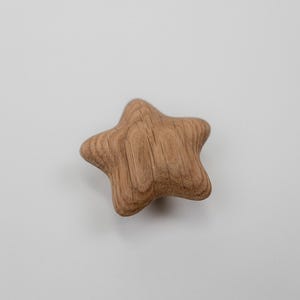Puede incluir: Objeto en forma de estrella de madera con un acabado natural de vetas. La estrella tiene cinco puntas redondeadas y está sobre un fondo blanco liso. Este artículo decorativo podría usarse como tirador de cajón o como acento decorativo.