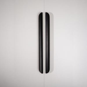 May include: Two black cabinet pulls with rounded ends are vertically aligned against a white background. The pulls are long and slender, with a smooth, matte finish. The design is minimalist and modern, suitable for various furniture styles.