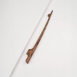May include: A single, long, wooden cabinet pull with a natural wood grain finish. The handle has a curved design with a wider base and a tapered end. The pull is set against a white background.