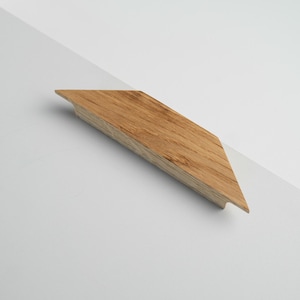 May include: Angled wooden furniture handle with a natural wood grain finish. The handle is a light brown colour and has a geometric, angular shape. The handle is designed to be mounted on a cabinet or drawer. The handle is approximately 25 cm long.