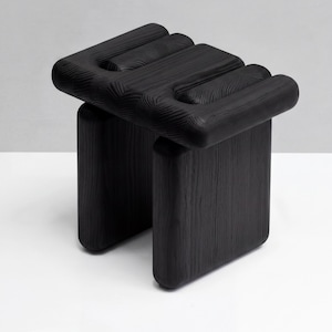May include: A black wooden stool featuring a rectangular seat with three distinct, rounded, parallel raised sections. The stool is supported by two thick, solid, rectangular legs with rounded corners, showcasing a visible wood grain texture throughout.