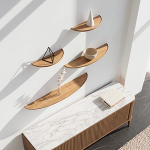 Puede incluir: Cuatro estantes de pared de madera en forma de semicírculo montados en una disposición escalonada. Los estantes presentan un acabado de veta de madera natural. Un cono blanco, un cuenco pequeño y un jarrón de vidrio con una planta seca están colocados sobre los estantes. Debajo hay una consola larga con una superficie de mármol blanco y listones de madera verticales.