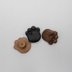 May include: Three wooden paw-shaped cabinet knobs in varying shades of brown and black. One knob is light brown with a screw on the back, another is solid black, and the third is a darker brown. These decorative knobs add a unique touch to furniture.