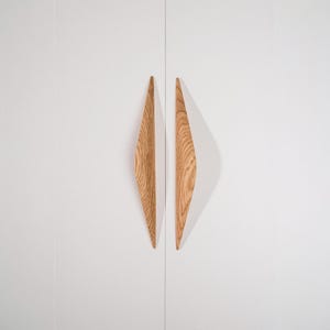 Op de afbeelding: Twee diamanten houten kastgrepen, gemonteerd op een witte kastdeur. De grepen zijn gemaakt van lichtgekleurd hout met zichtbare nerf en werpen een schaduw op de deur.
