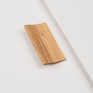 Puede incluir: Un tirador de mueble rectangular de madera con un acabado de veta natural en color marrón claro. El tirador presenta un perfil ligeramente curvado y cóncavo, montado sobre una superficie blanca plana, mostrando un diseño simple y limpio.